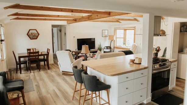 A modern kitchen with a central island, wooden beams, and a dining area with a table and chairs. - Charming IU Cottage: Near Campus, Sleeps 8! (Bloomington)