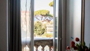 A room with a large window, a balcony with a view of trees and buildings, and a vase with flowers on a table. - Agora Apartments   Sea View Apartment (Sorrento)