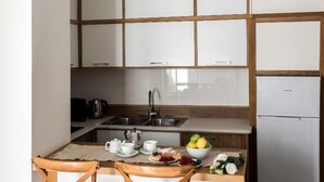 A compact kitchen with white cabinets, a stainless steel sink, and a small refrigerator. There are two wooden bar stools and a table with a kettle, cups, and a bowl of fruit.