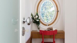 A corner room with a wooden desk, a red chair, a round window with a view of greenery, and a door with a glass panel.
