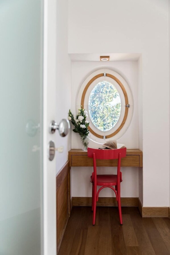 A corner room with a wooden desk, a red chair, a round window with a view of greenery, and a door with a glass panel.