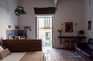 Ein gemütliches Zimmer mit einem Bett, einem Schreibtisch, einem Sessel und einer Couch. Ein Fenster bietet Blick auf einen Balkon und ein Gebäude.