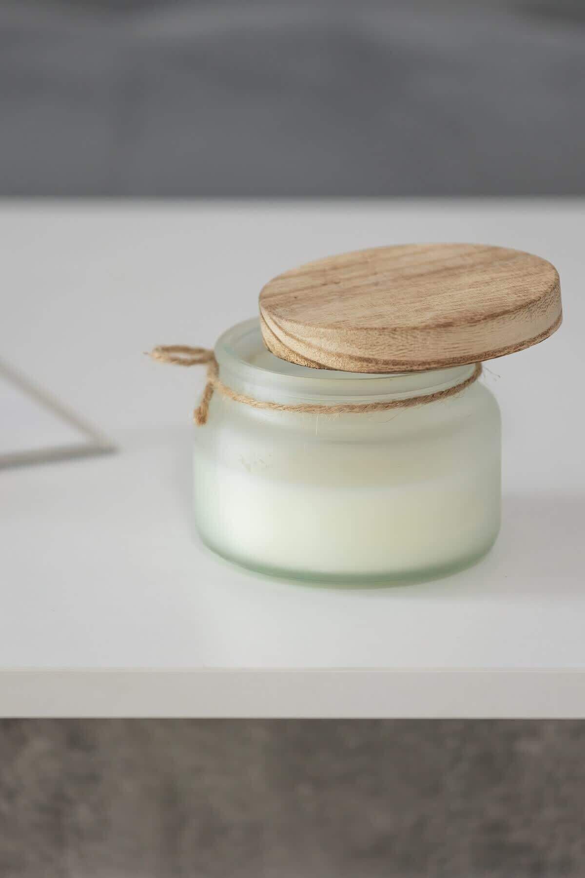 Un bocal en verre transparent avec un couvercle en bois et un morceau de ficelle, posé sur une surface blanche.