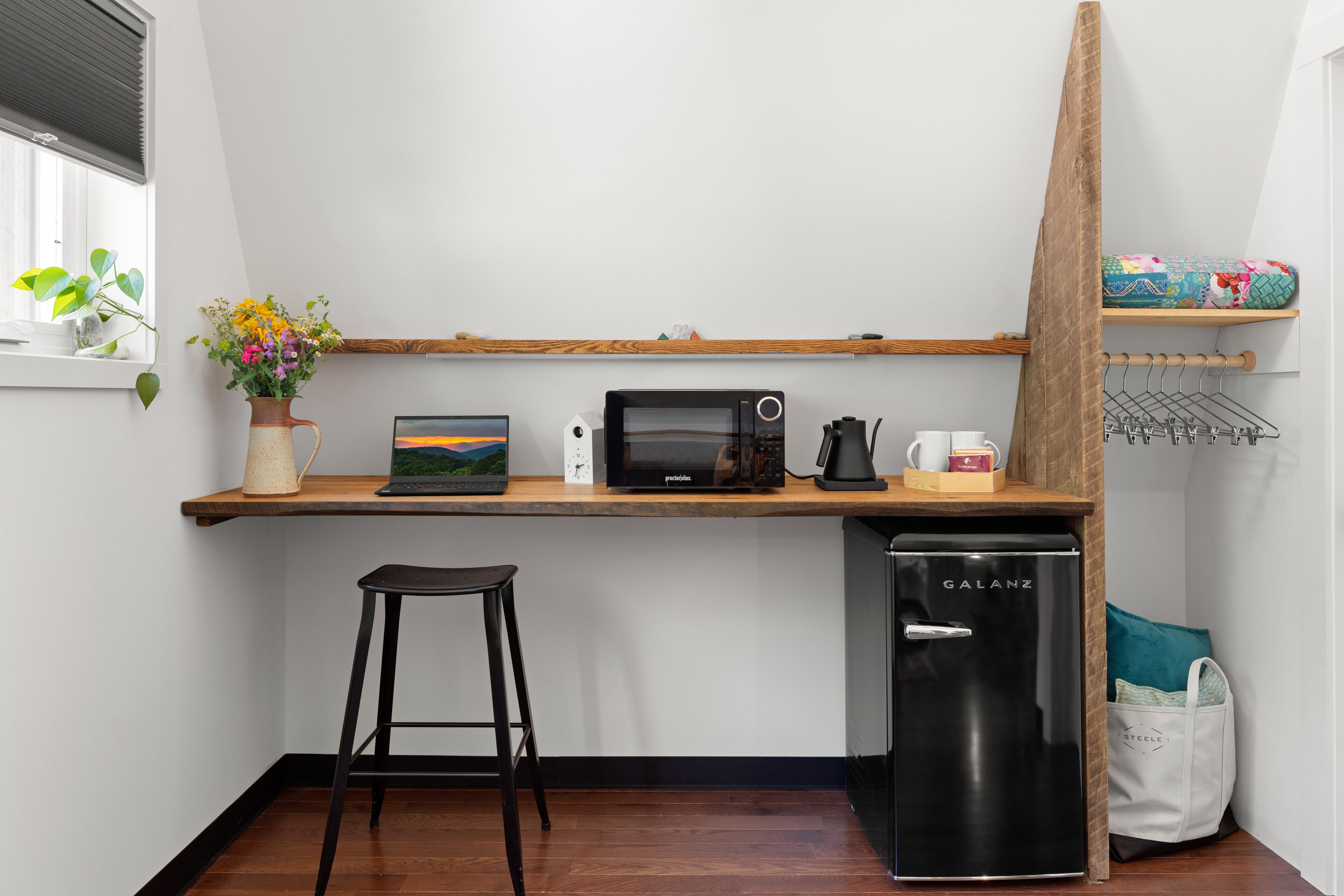 A compact kitchen with a microwave, coffee maker, and a refrigerator. A wooden shelf above the counter holds a plant and a painting.