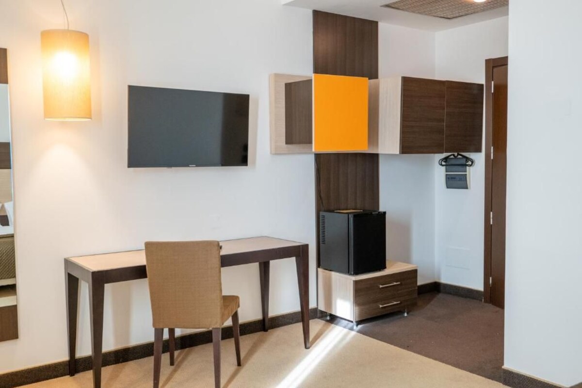 Quarto de hotel compacto com escrivaninha, cadeira, TV de tela plana, guarda-roupa e um pequeno frigorífico.