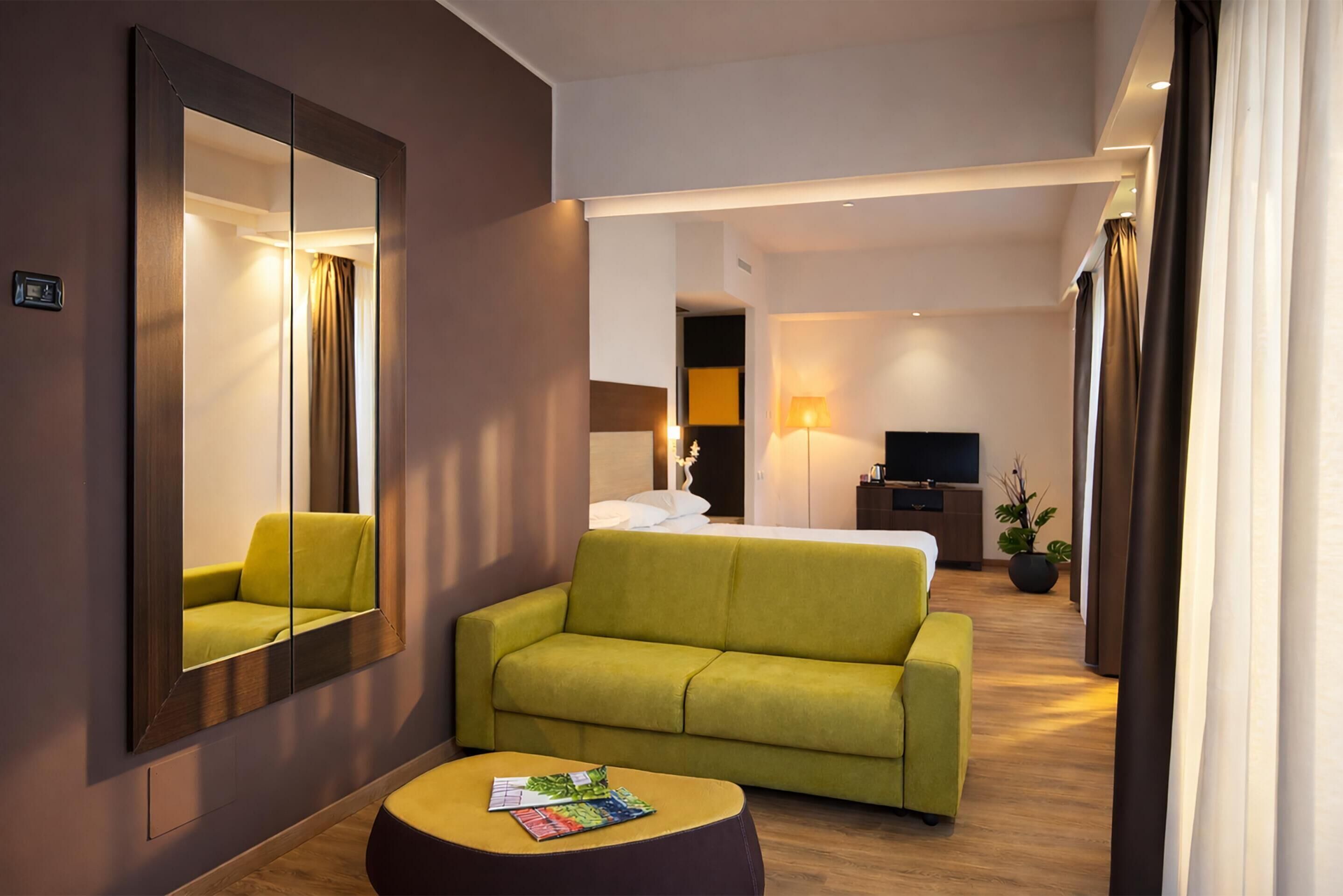 Una habitación de hotel con un sofá verde, un reposapiés pequeño y redondo, un espejo grande, una cama con ropa blanca, un televisor y una planta en maceta.