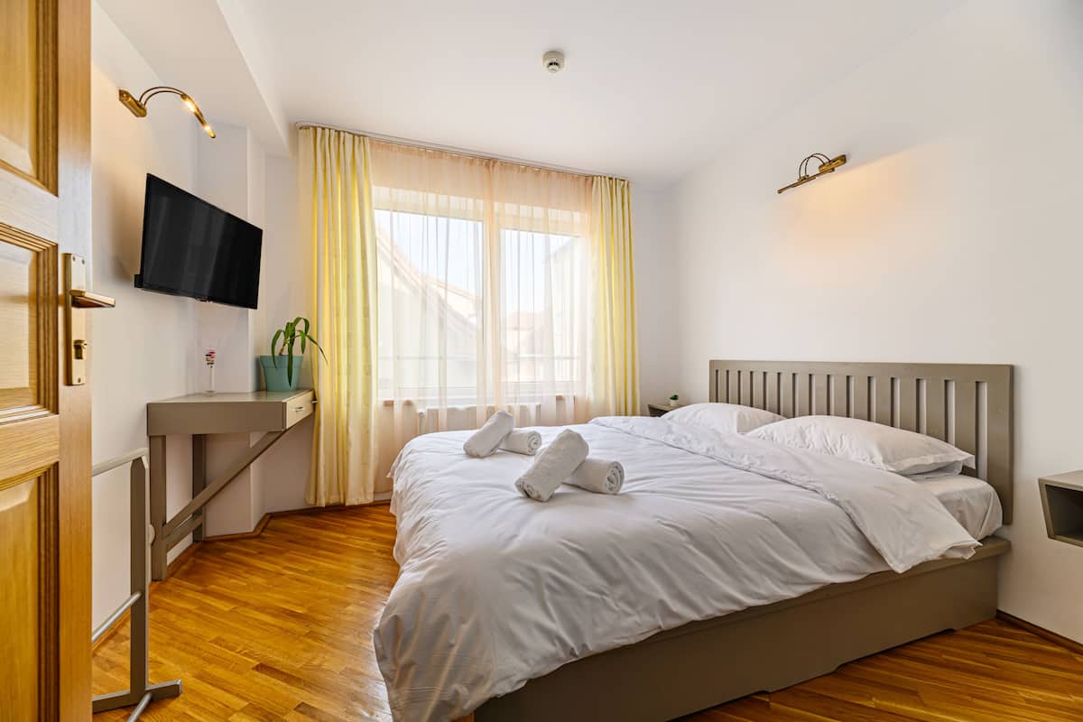 A hotel room with a bed, a TV mounted on the wall, a desk, and a window with curtains.