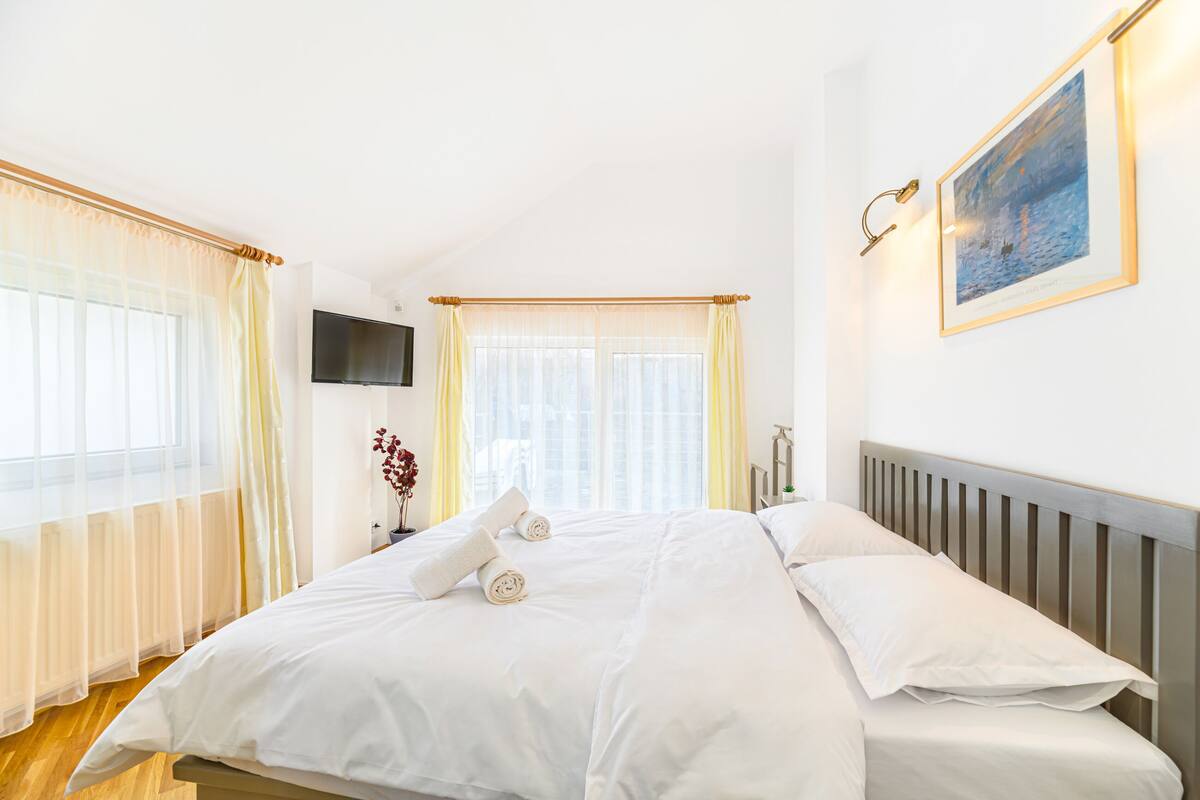 A neatly made bed with white linens and two rolled towels, a flat-screen TV mounted on the wall, and a framed picture above the bed.