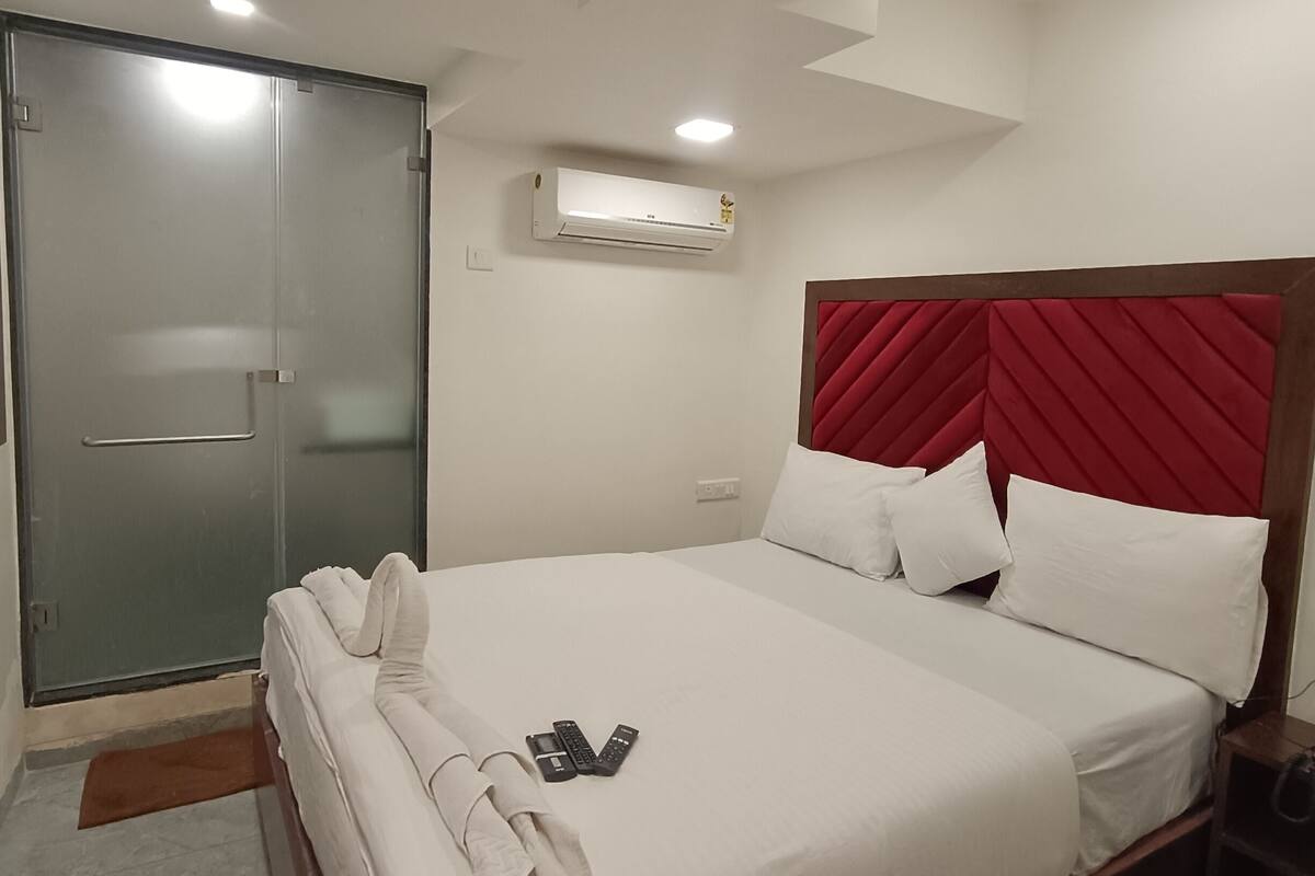 A hotel room with a bed, a red headboard, an air conditioning unit, a glass door, and a bedside table.