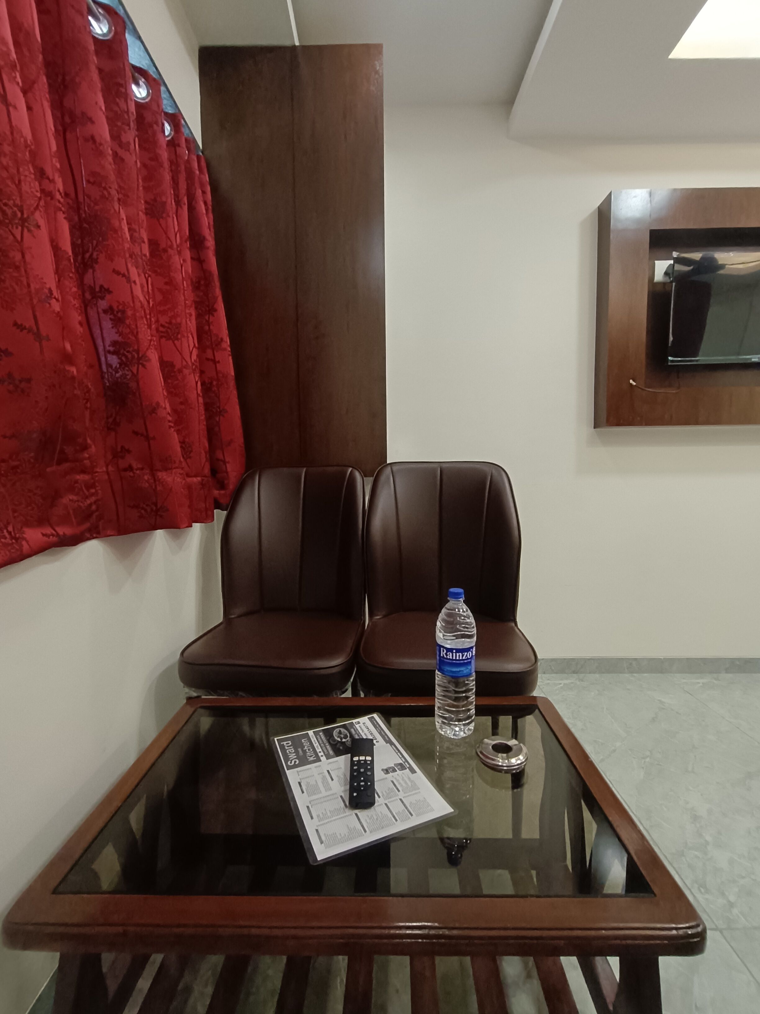 Quarto com duas poltronas de couro, uma mesa de vidro com um controle remoto e um jornal, uma garrafa de água e uma TV fixada na parede.