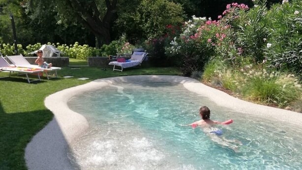 Une piscine où un enfant nage, entourée d’un jardin agrémenté de chaises longues et de verdure.