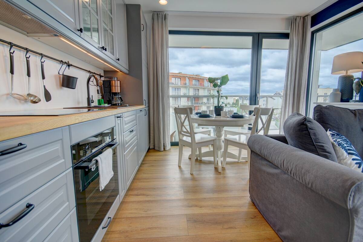 A modern kitchen with a dining area, large windows, and a grey sofa.