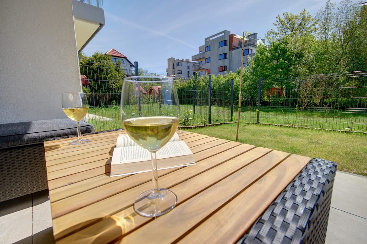 A wooden table with two glasses of white wine, a book, and a view of a residential area with buildings and greenery.