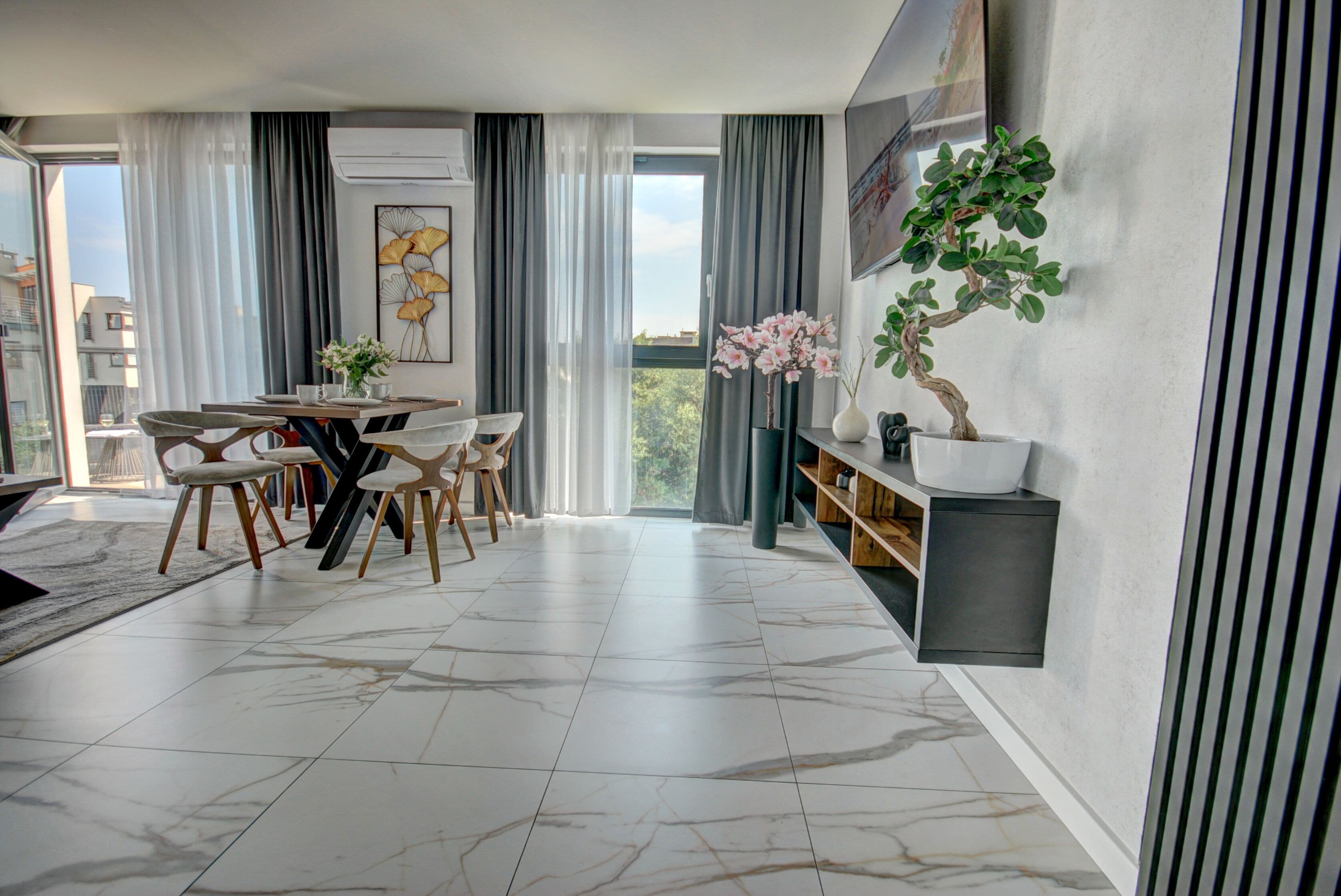 A modern dining area with a marble floor, a round table, and chairs. There is a large window with curtains, a wall-mounted air conditioner, and a potted plant on a shelf.