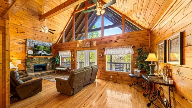 A spacious log cabin interior with a fireplace, a stone wall, a large sofa, a dining table, and a ceiling fan. - Smoky Mountain Getaway: Leave the World Behind (Pigeon Forge)