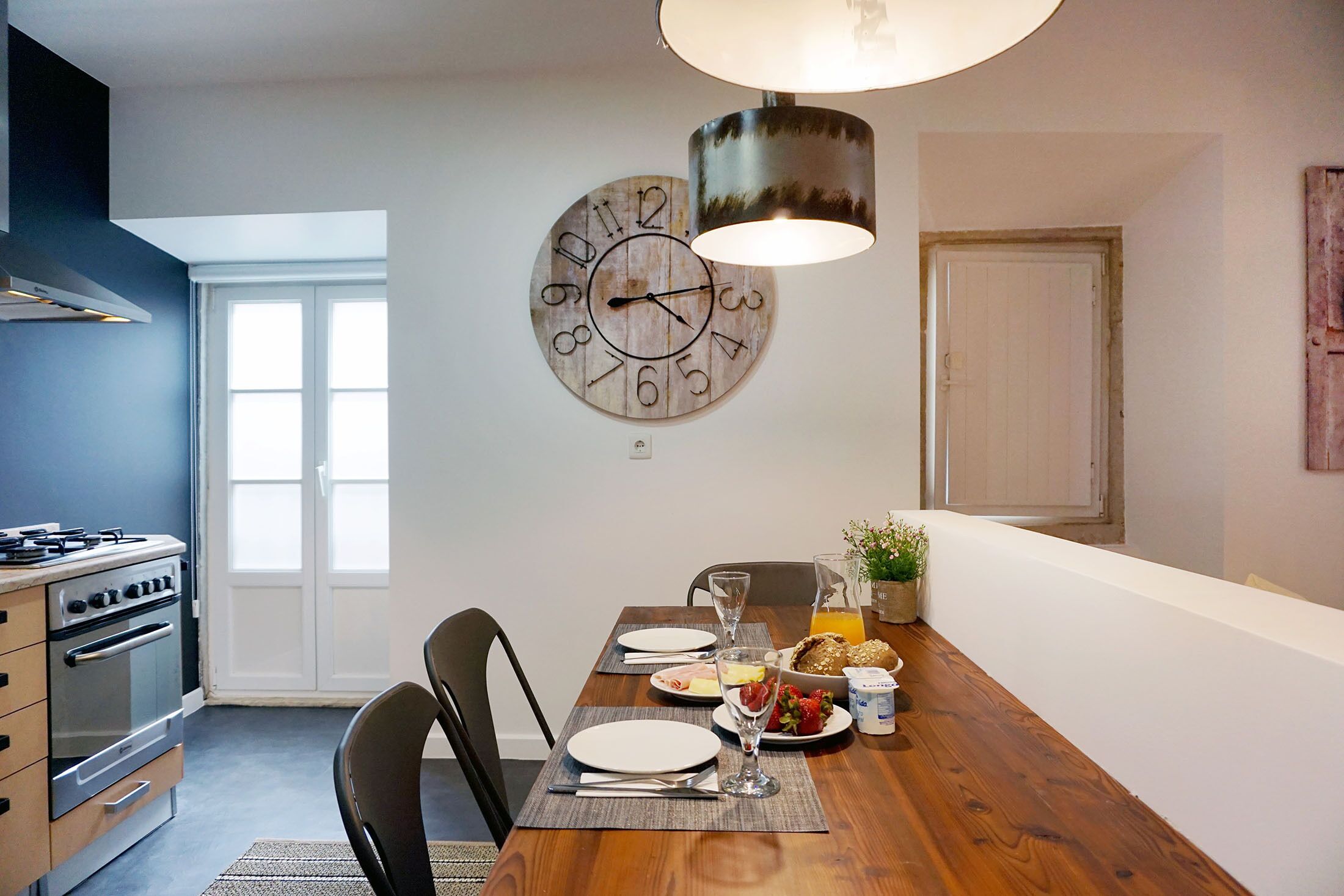 Un comedor con una mesa de madera dispuesta para dos personas, un reloj en la pared y una cocina al fondo.