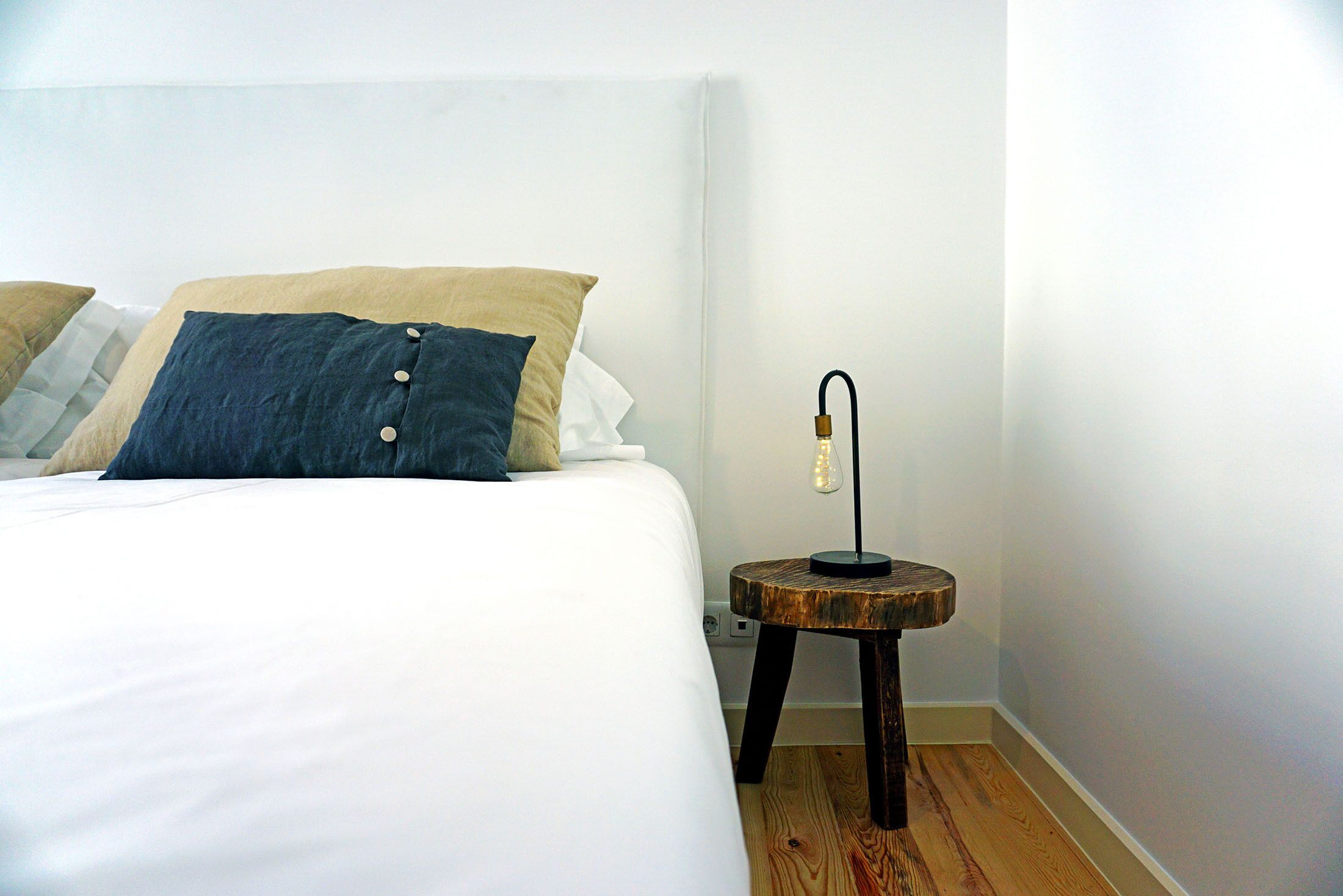 A neatly made bed with white and dark blue pillows, a small wooden nightstand with a lamp, and a wooden floor.