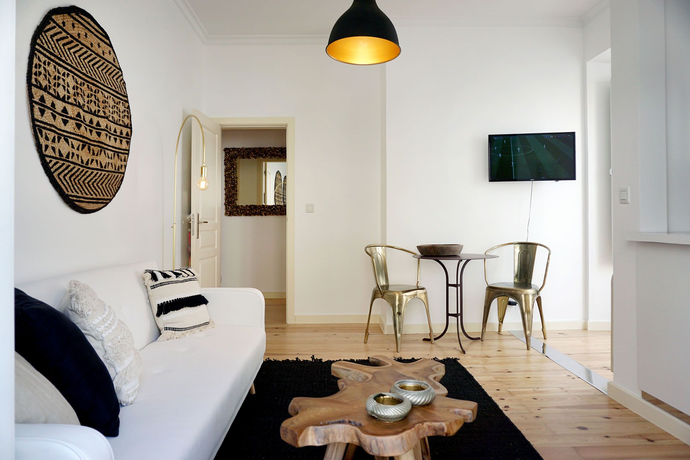A modern living room with a white sofa, a wooden coffee table, and two chairs. A television is mounted on the wall, and a decorative wall hanging is visible.