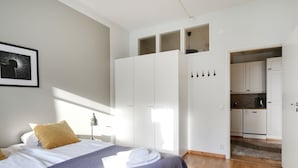 A bedroom with a bed, a wardrobe, a kitchen area, and a framed picture on the wall. - Kotimaailma Aparthotel Kamppi - 2BR (Helsinki)