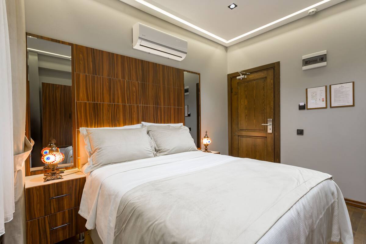 A neatly made bed with a wooden headboard, a bedside lamp, and a wall-mounted air conditioner.