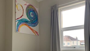 A bedroom with a white quilted bed, a colorful abstract painting on the wall, and a window with grey curtains. - Central 2 bed flat. Free parking (Off-street) (Bristol)