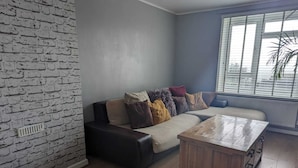 A living room with a brick wall, a sofa with multiple cushions, a wooden coffee table, and a window with blinds. - Seaview/downs National Park & Free Parking (Brighton)