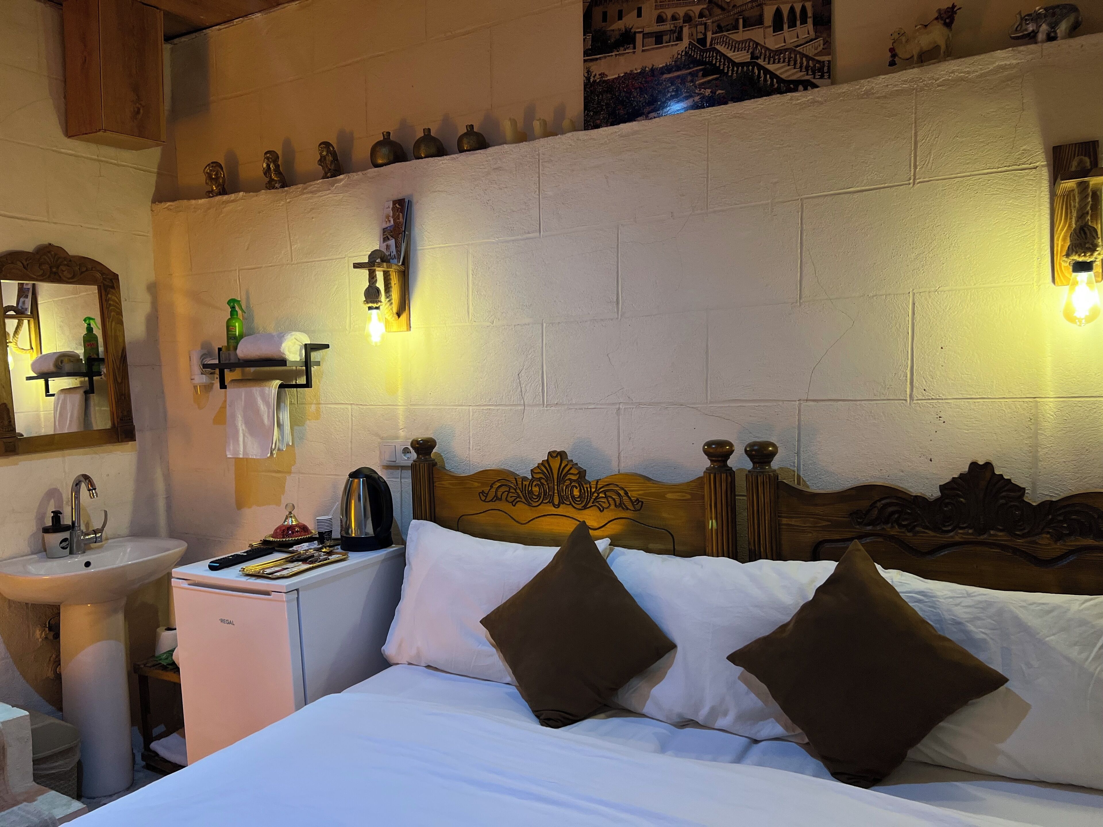 A hotel room with a wooden bed, white bedding, a small bedside table with toiletries, a wall-mounted shelf with decorative items, and a white sink.