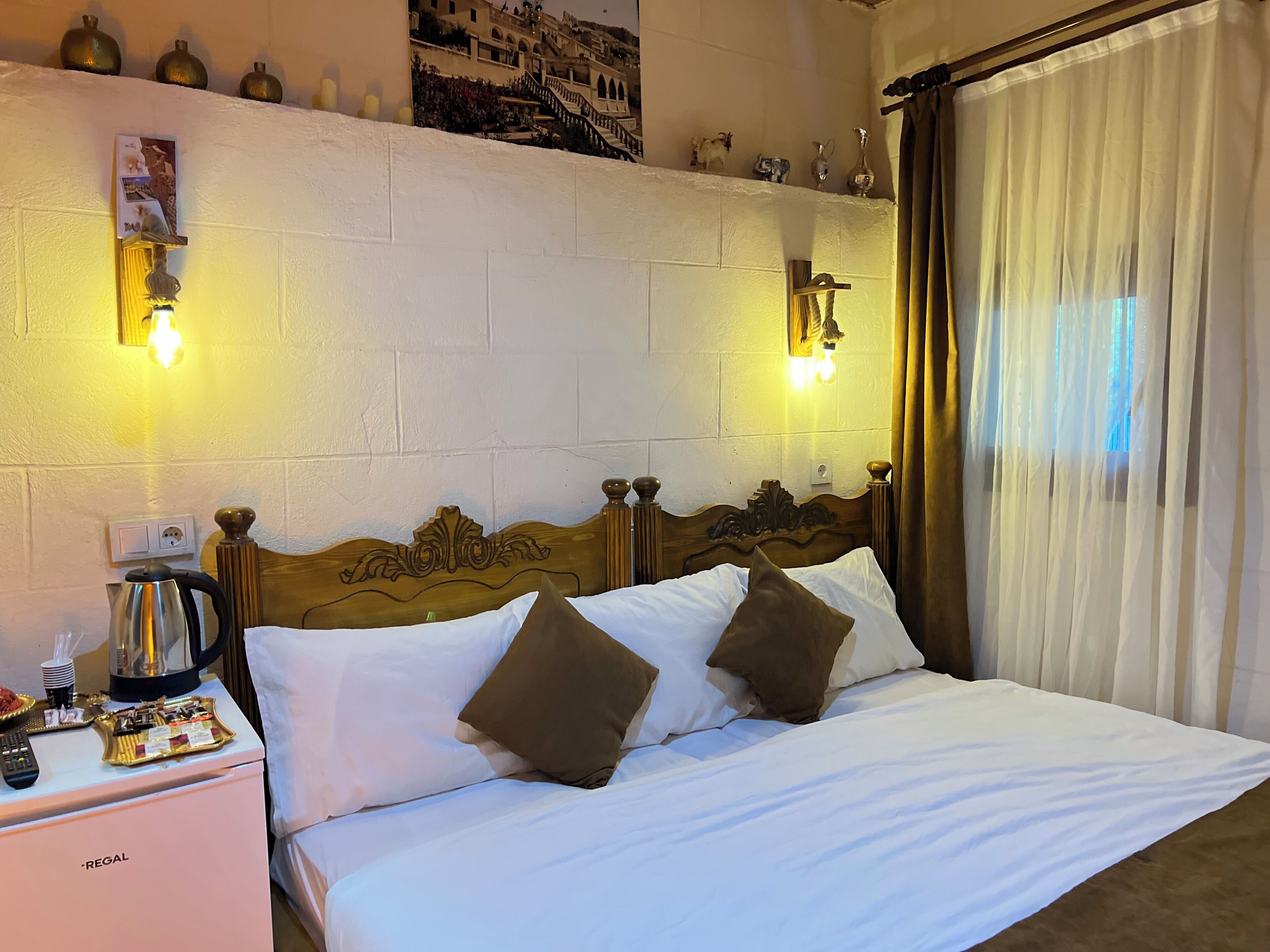 A hotel room with a wooden headboard, two pillows, a bedside table with a kettle, and a nightstand.