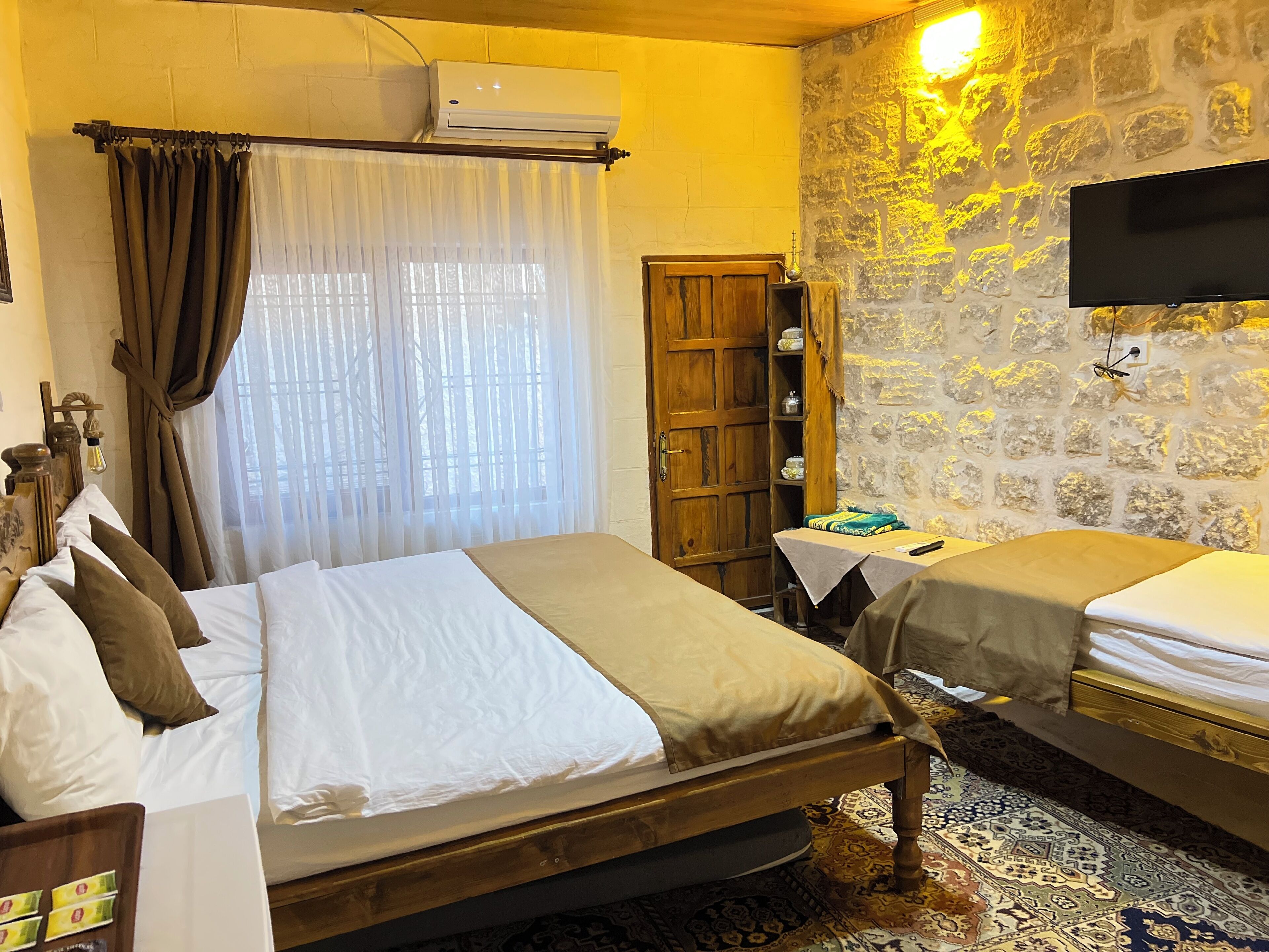A hotel room with two beds, a wooden wardrobe, a small table, an air conditioning unit, and a television on the wall.