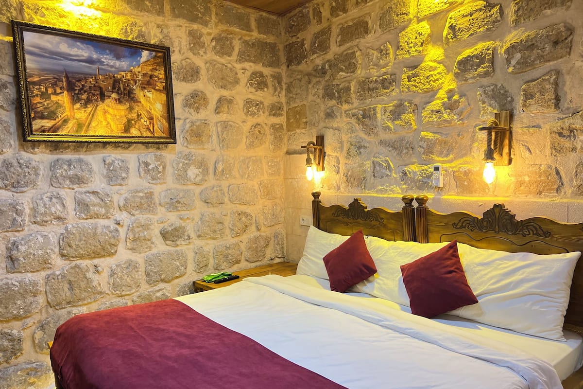 Quarto com parede de pedra, cama com roupa de cama branca e vinho, e pintura de uma paisagem urbana.