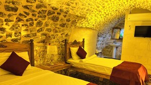Una habitación con dos camas, paredes de piedra, cabecero de madera, una mesita de noche, una lámpara y un baño visible a través de un arco.