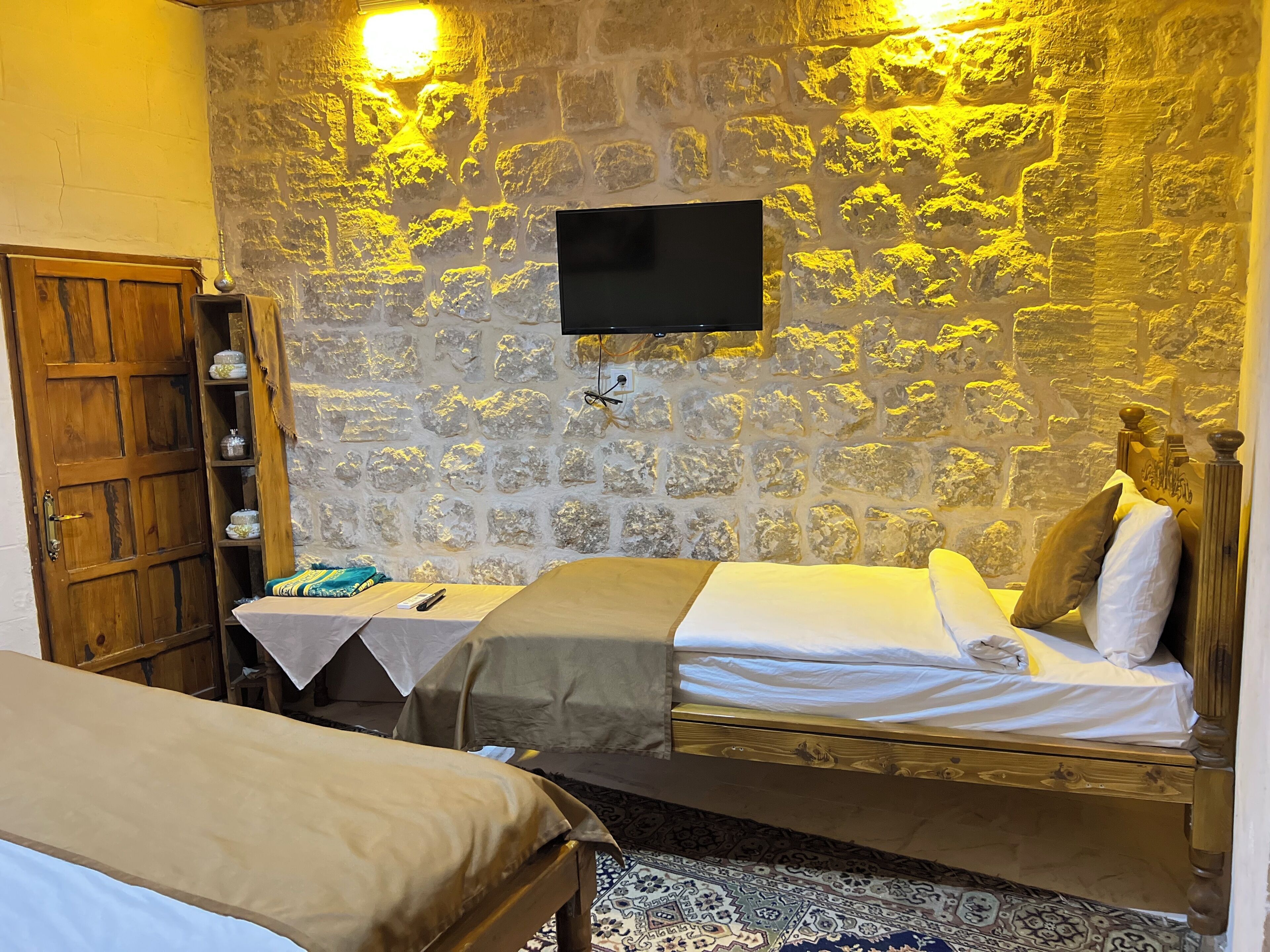 Une pièce avec un mur en pierre, un téléviseur fixé au-dessus d’un lit, une armoire en bois et une table de chevet avec une lampe.