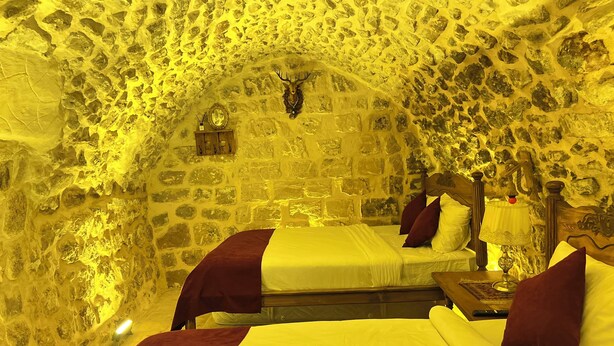 Una habitación con paredes de piedra, dos camas con ropa de cama roja y blanca, un cabecero de madera, una mesita de noche con lámpara y un reloj en la pared.