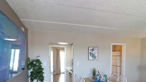 Ett rum med en platt-TV, ett matbord med stolar, en krukväxt och ett inramat konstverk på väggen.