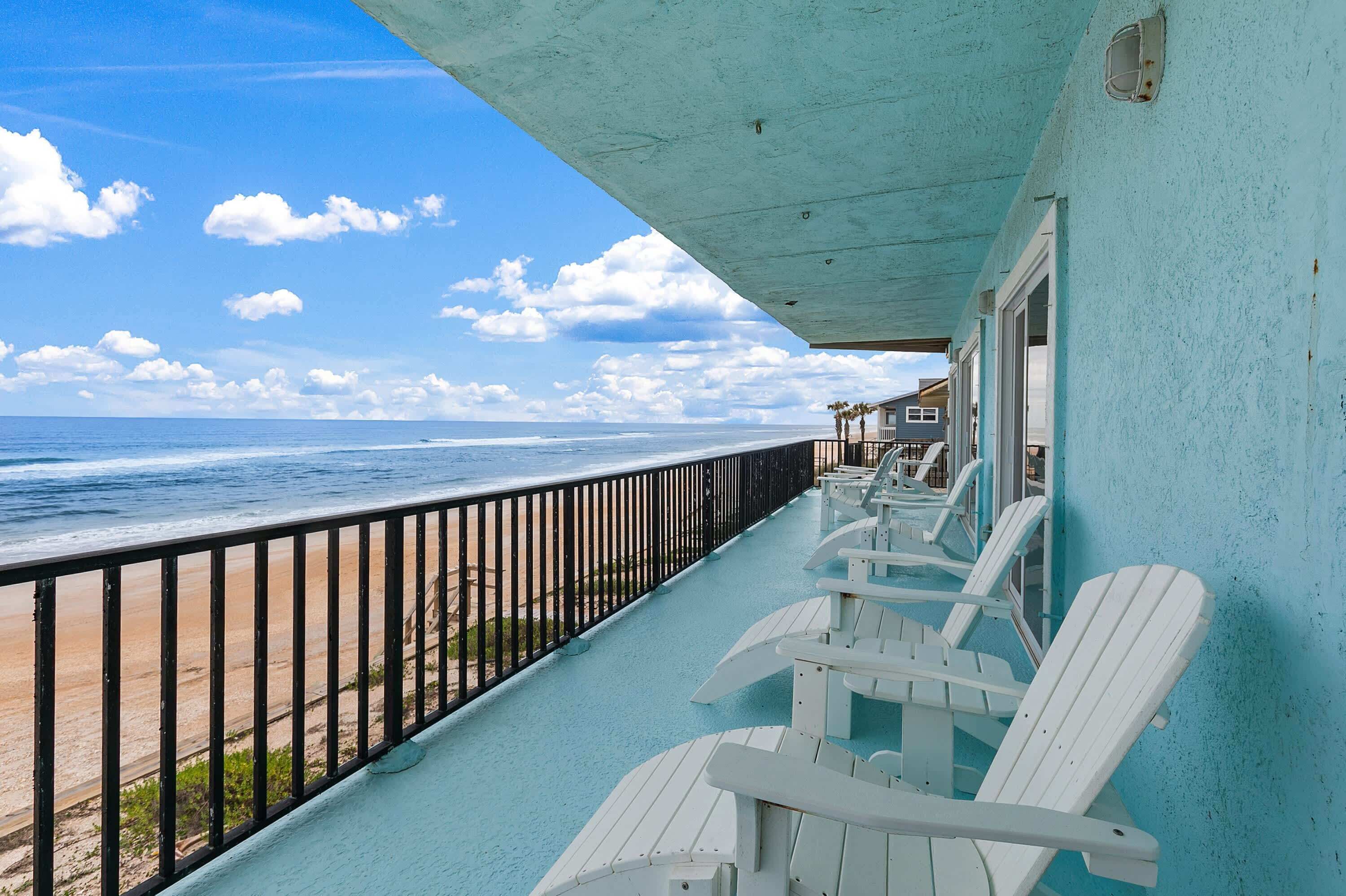 A balcony with white Adirondack chairs, a view of the beach, and a building with a light blue exterior.