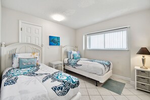 A bedroom with two beds, a window with blinds, a door, a small table, and a lamp.