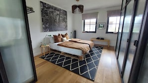 A bedroom with a bed, bedside tables, a hanging light fixture, a large cityscape painting, and a window with a view.