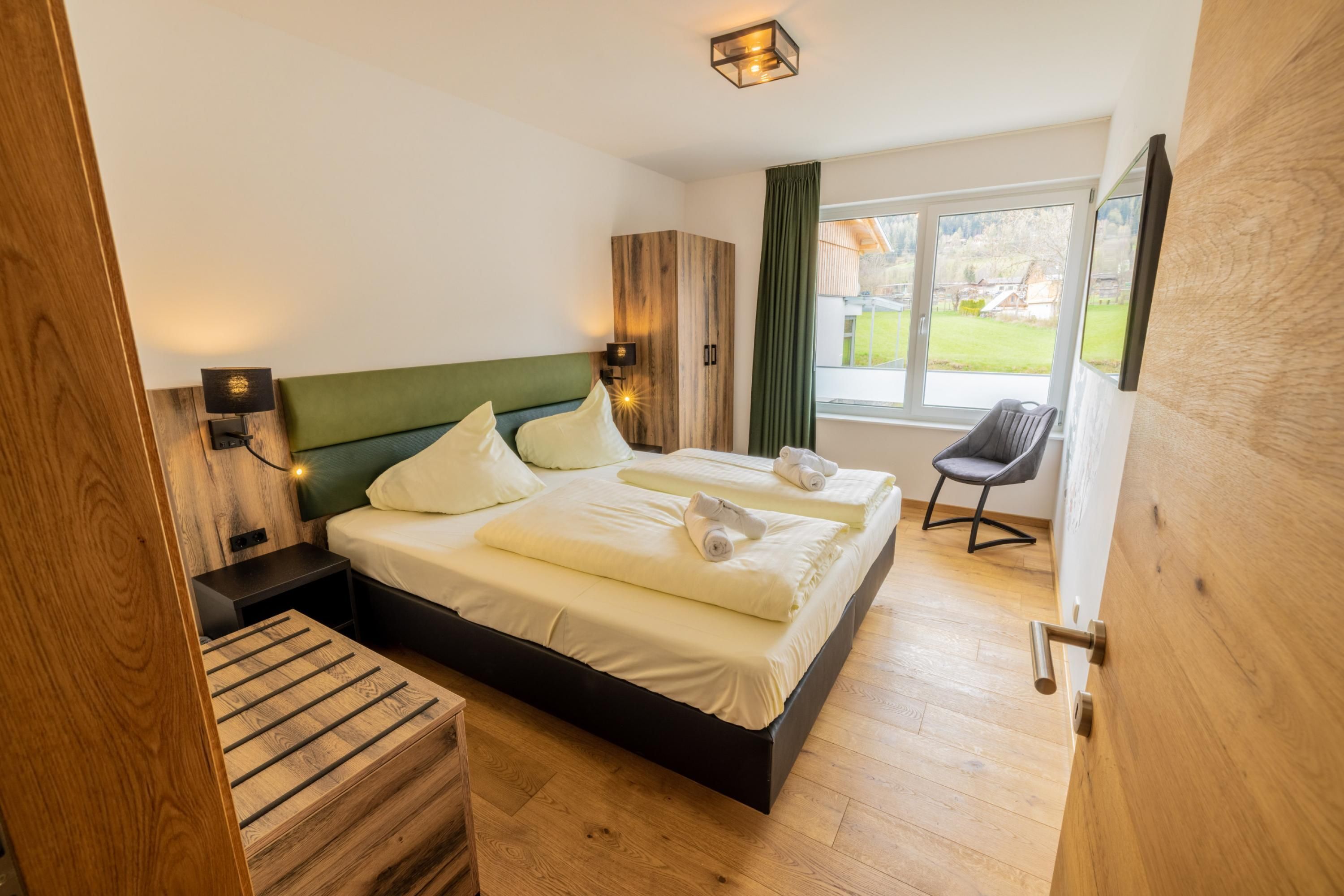 A modern bedroom with a wooden bed frame, two bedside tables, a chair, and a large window with a view of the outdoors.