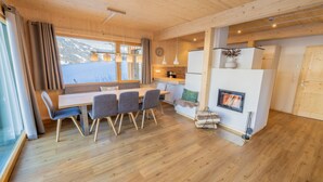 A modern interior with a wooden floor, a dining area with a table and chairs, a kitchen with a stove, and a large window with curtains.