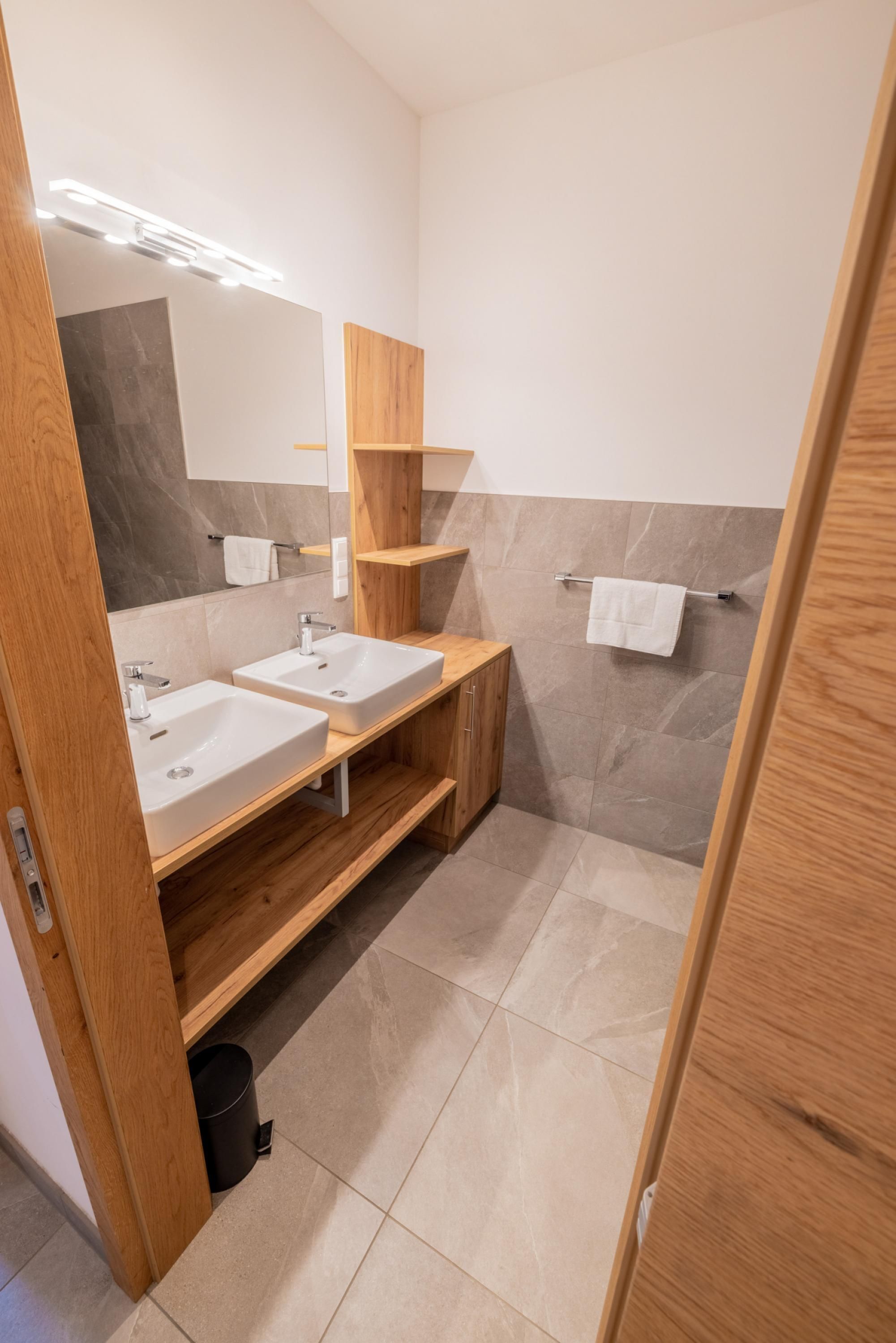 Un baño moderno con dos lavamanos, estantes de madera, un espejo y un perchero para toallas.