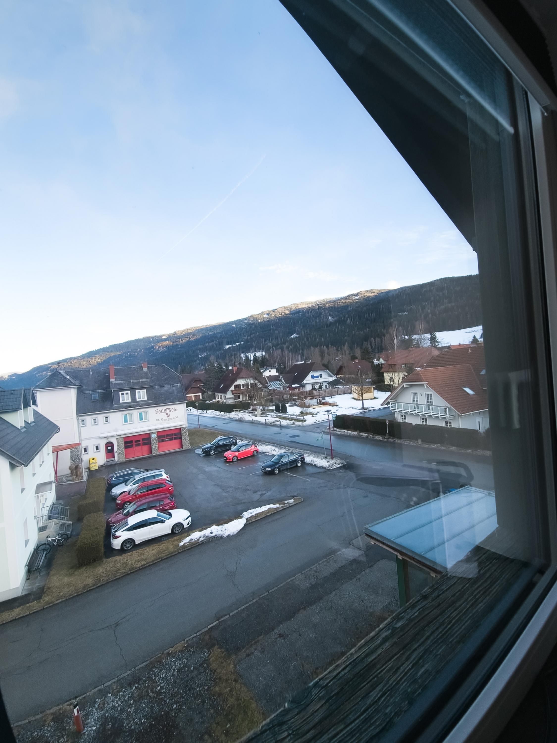 A parking lot with several cars, a building with a red garage door, and a view of a hilly landscape in the background.