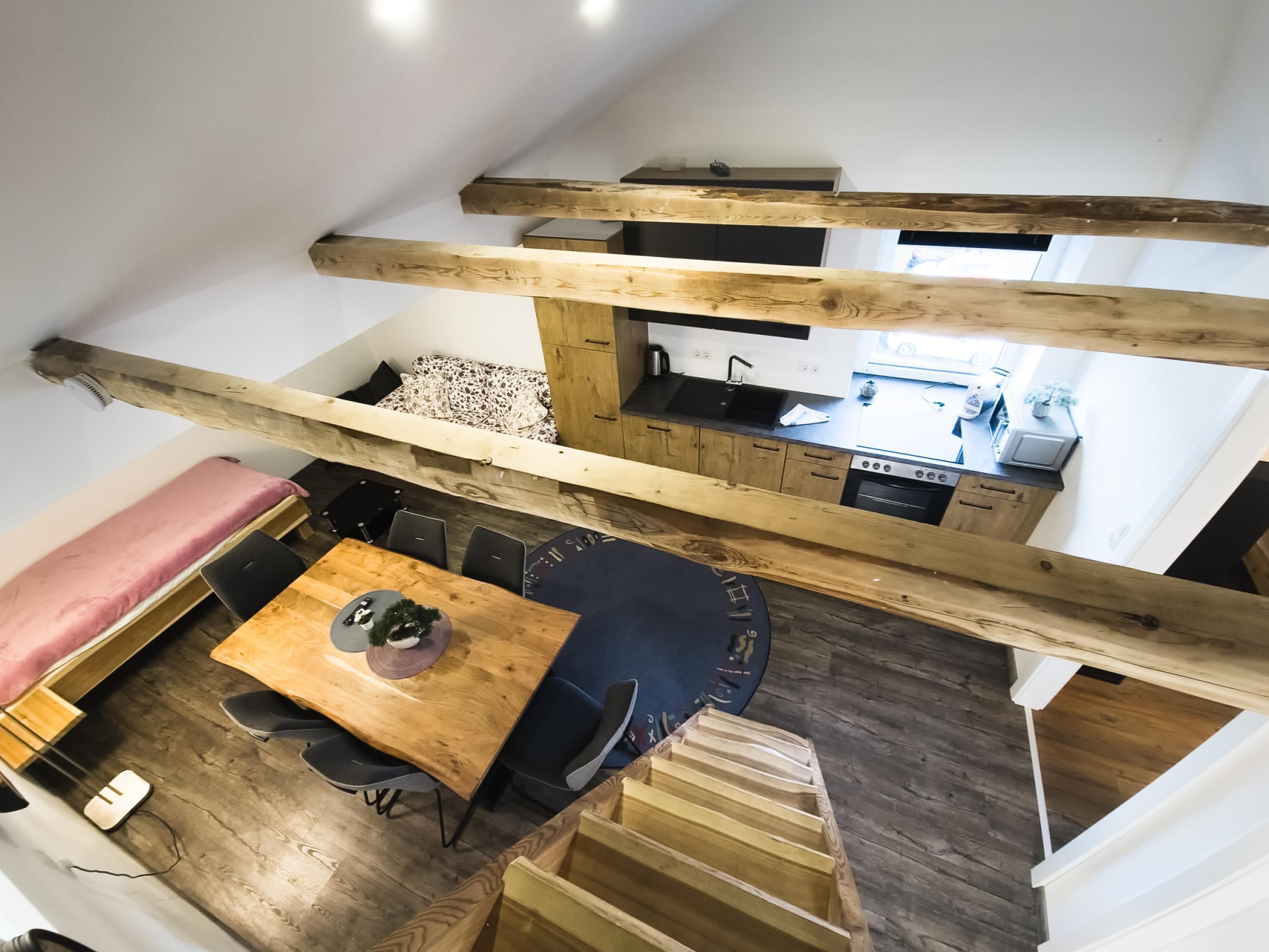 Et loft-studie med træloft, spiseplads med bord og stole, et tekøkken og en seng med et mønstret sengetæppe.