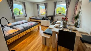 A compact room with two bunk beds, a sofa, a dining table set for four, and large windows with curtains.