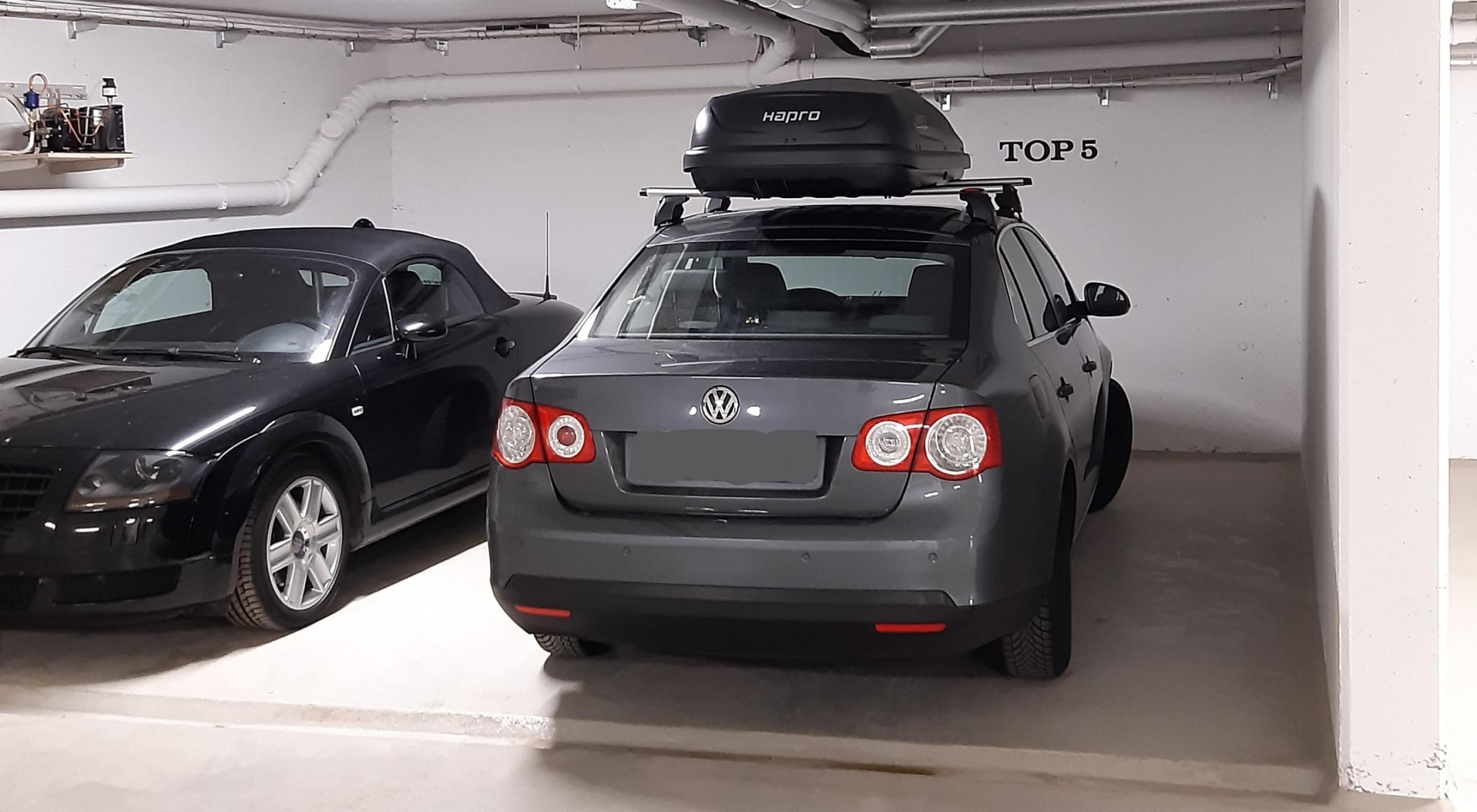 Twee auto's staan geparkeerd in een garage met een witte muur en een bord waarop 'TOP 5' staat.