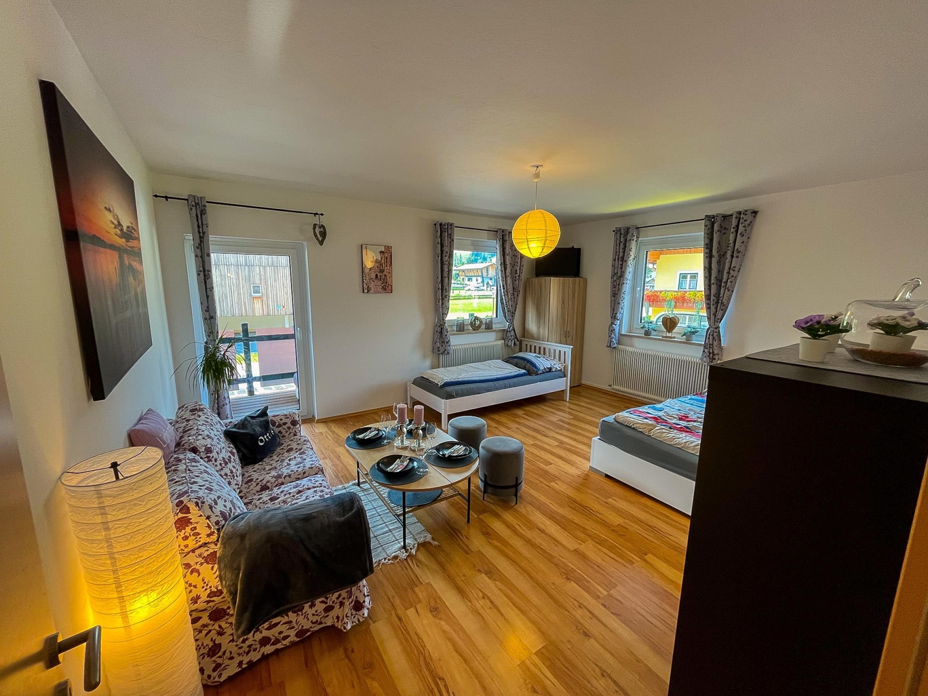 A cozy living room with a sofa, a small dining table, and a bed. There are windows with curtains, a hanging light fixture, and a painting on the wall.