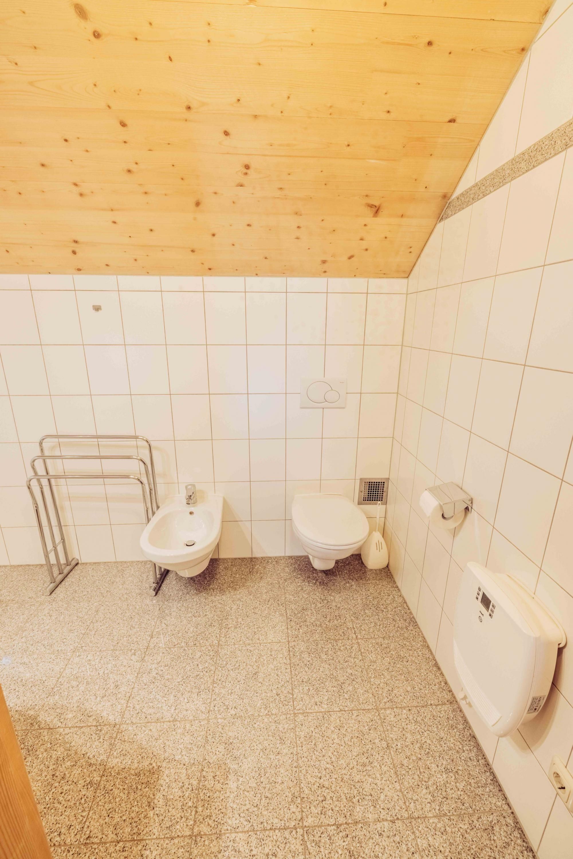 Un baño pequeño y limpio con techo de madera, paredes de azulejos blancos y suelo de baldosas. Incluye un lavamanos, un inodoro y un bidé.