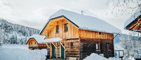 A wooden cabin with a snow-covered roof, surrounded by snow and trees.