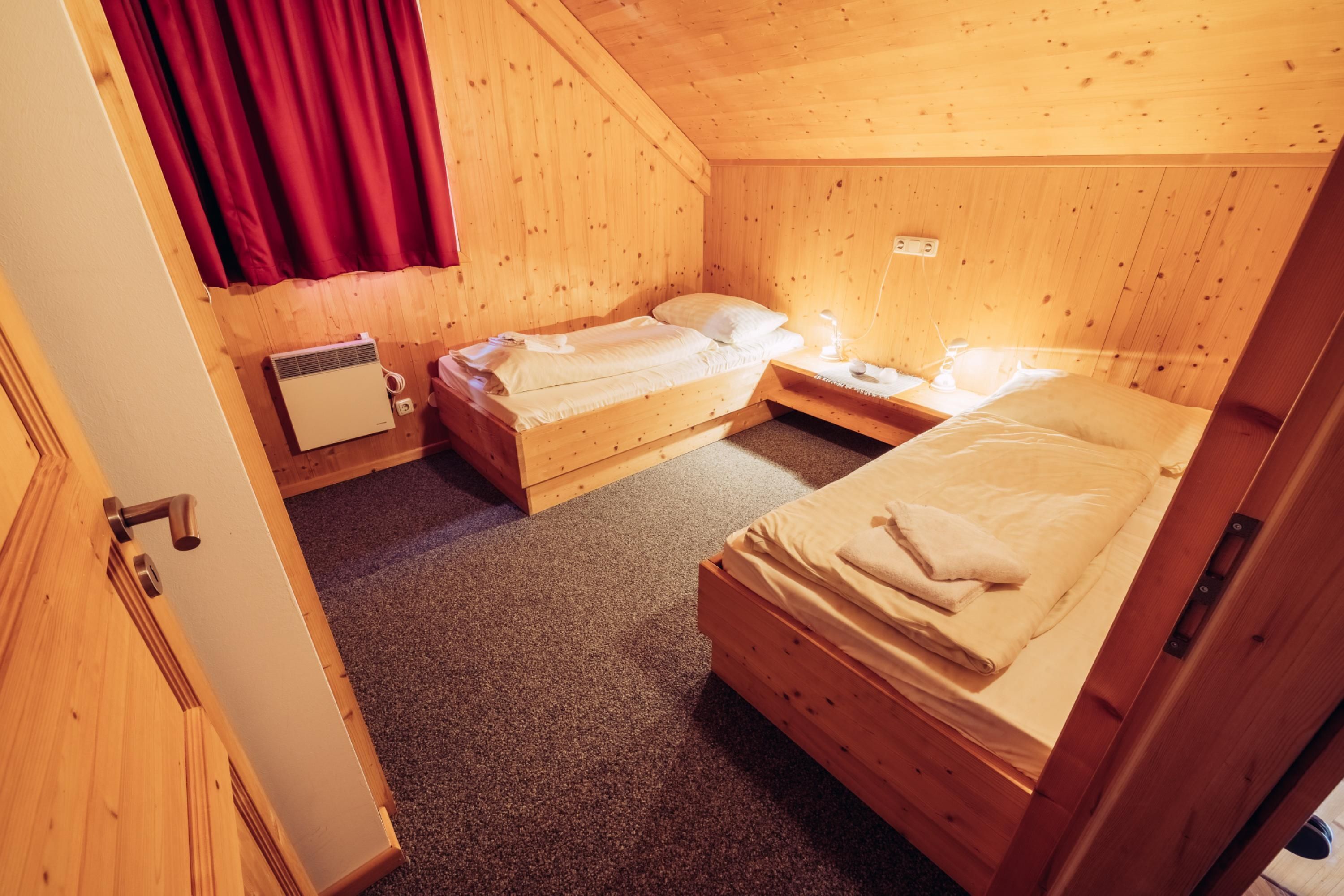 A small, cozy room with two wooden beds, a red curtain, a wall-mounted heater, and a small table with a lamp.
