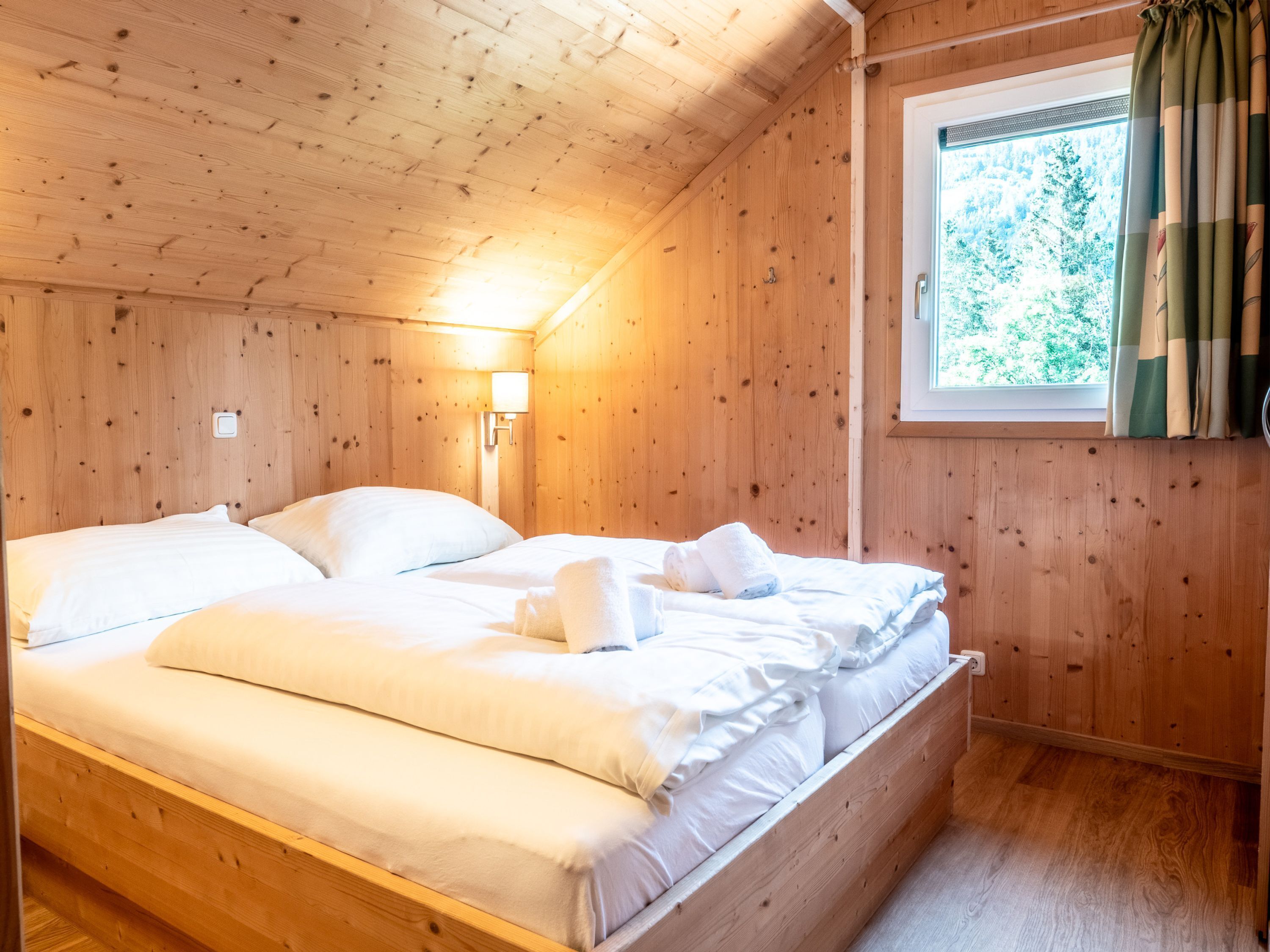 A wooden cabin bedroom with a bed, a nightstand, a wall-mounted lamp, and a window with curtains.
