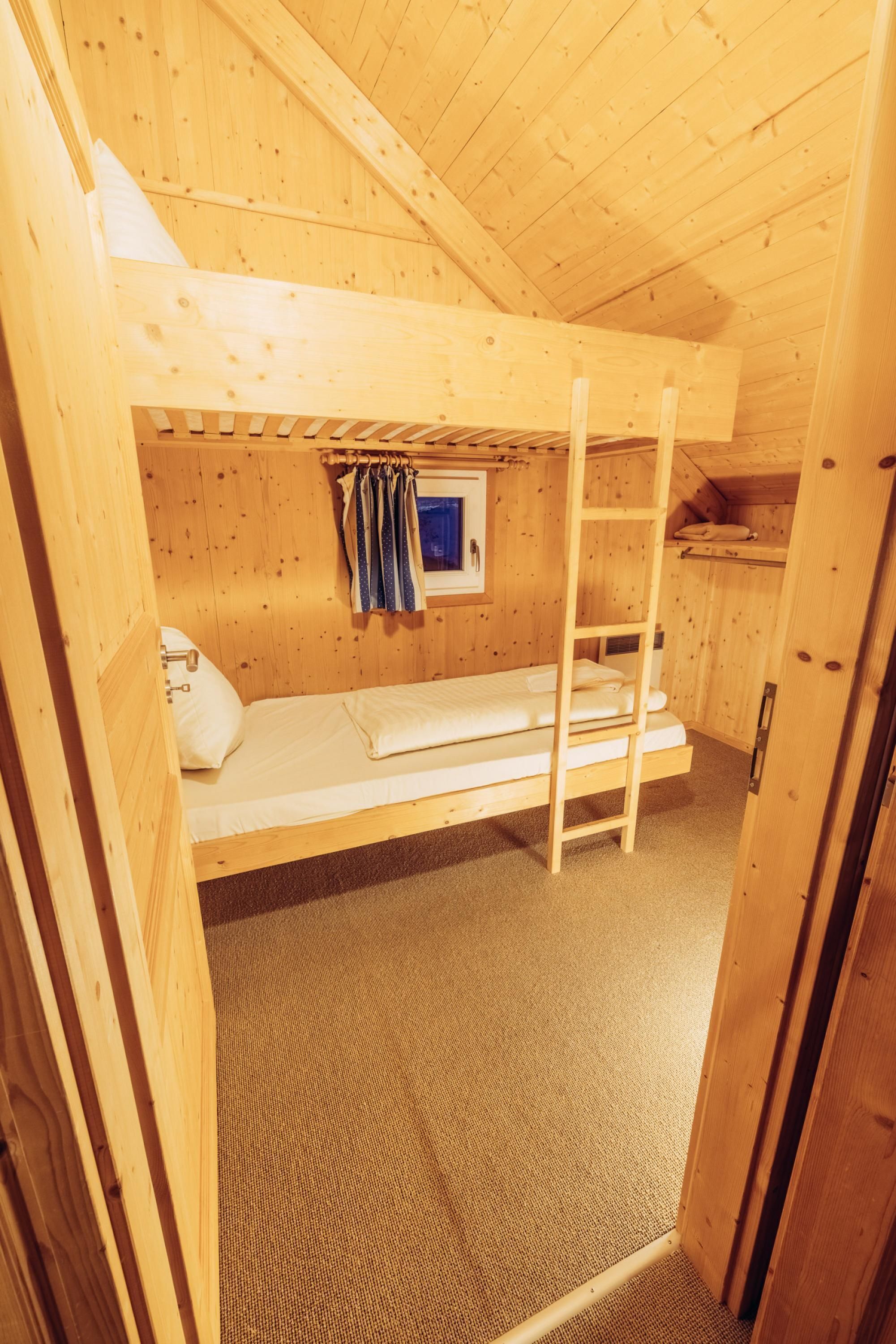 A small, wooden cabin room with a bunk bed, a window with curtains, and a ladder.